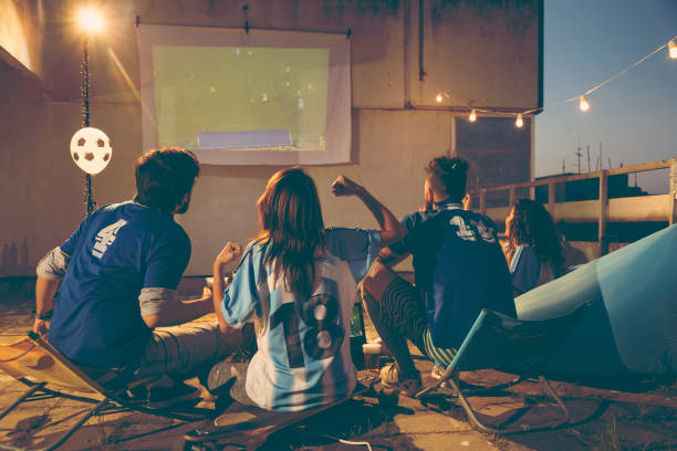 Group of young friends watching a football match on a building rooftop terrace, drinking beer and cheering for their team