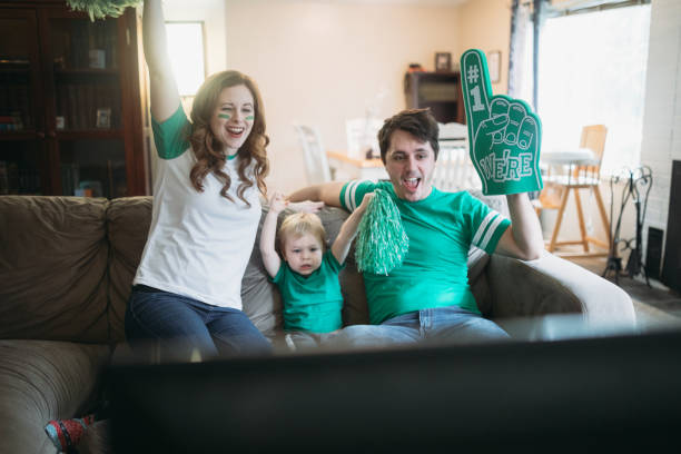 A happy family with a young child have fun goofing off while watching a sports game on the television.  They wear their team color and celebrate a win!